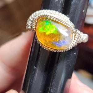 May include: A silver ring featuring a large, oval-shaped opal. The opal displays vibrant colors, including yellow, green, and blue, with a shimmering, iridescent effect. The ring's band has a textured, braided design.