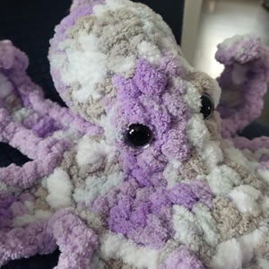 Könnte beinhalten: Ein gehäkeltes Plüsch-Kraken-Spielzeug aus lila, weißem und grauem Garn. Der Krake hat ein großes schwarzes Auge.