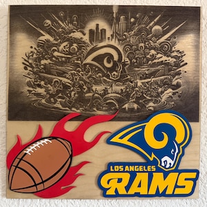 May include: A wooden plaque with a laser-etched image of a football scene with a cityscape in the background. The plaque has a red and yellow flame design with a brown football and a blue and yellow Los Angeles Rams logo.