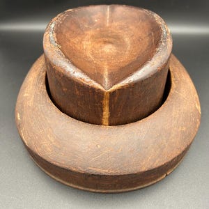 May include: A vintage wooden hat block for making hats. The block is made of two pieces, a top and a bottom. The top is a rounded shape and the bottom is a flat circle. The block is brown and has a worn look.