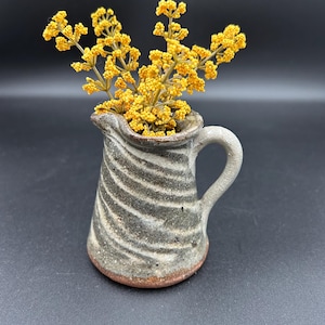 May include: A small, handmade ceramic pitcher with a gray and white striped pattern, holding a bouquet of bright yellow flowers. The pitcher has a small handle and a brown base, set against a dark background.