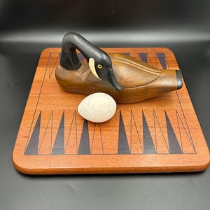 May include: A wooden sculpture of a goose with a black head and a brown body, resting on a wooden backgammon board. A small, white, oval stone sits in front of the goose. The board is reddish-brown with black and white triangular inlays.