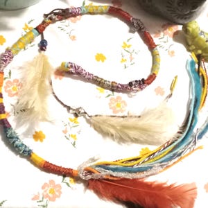 May include: A colorful braided hair wrap with feathers. The wrap is made of multiple strands of yarn in shades of red, orange, yellow, blue, and purple. It has three feathers attached, one white and two orange.