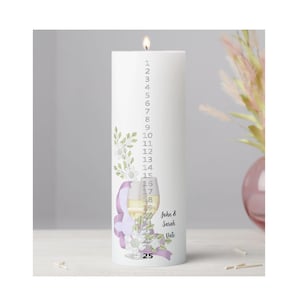 May include: A white cylindrical pillar candle with a lit wick, featuring a vertical countdown from 1 to 25. The design includes a printed illustration of a wine glass, green leaves, white flowers, a purple ribbon, and the text John & Sarah Date.