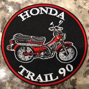 May include: A black and red embroidered patch with a white outline. The patch features a red Honda Trail 90 motorcycle with the words "Honda Trail 90" in white.