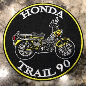 May include: A round black patch with a yellow border featuring a yellow and gray Honda Trail 90 motorcycle illustration. The words "HONDA" and "TRAIL 90" are embroidered in white, arching around the motorcycle image.