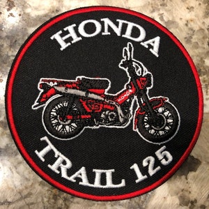 May include: A black and red embroidered patch with a white outline. The patch features a red Honda Trail 125 motorcycle with the words "Honda Trail 125" in white.
