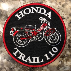 May include: A red and white embroidered patch with a black background. The patch features a red Honda Trail 110 motorcycle with the words "Honda Trail 110" in white.