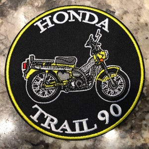 May include: Embroidered patch featuring a yellow and gray Honda Trail 90 motorcycle on a black background. The patch has a yellow border and the words "HONDA" and "TRAIL 90" in white.