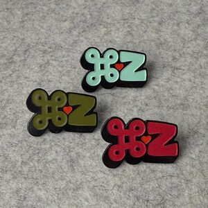May include: Three enamel pins with the letters "HZ" in a stylized font. The pins are in shades of green, red, and teal, each with a black outline and a small red heart. The pins are on a gray felt surface.