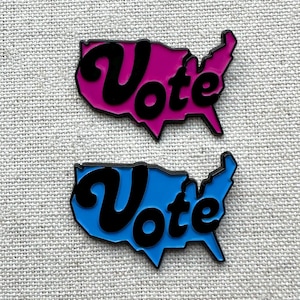 May include: Two enamel pins shaped like the United States of America with the word "Vote" in a stylized font. One pin is pink and the other is blue.