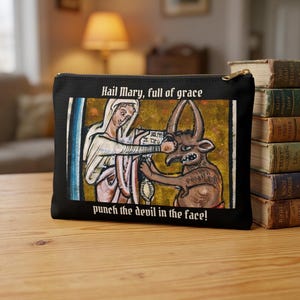 May include: Black zippered pouch featuring a medieval-style illustration of a woman punching a devil. The image includes the text "Hail Mary, full of grace" and "punch the devil in the face!" A stack of old books is visible in the background.