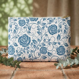 May include: A rectangular cosmetic bag with a white background and a repeating pattern of blue roses and vines. The bag has a gold zipper and is sitting on a brick surface.