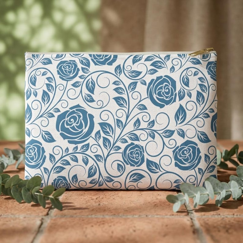 May include: A rectangular cosmetic bag with a white background and a repeating pattern of blue roses and vines. The bag has a gold zipper and is sitting on a brick surface.