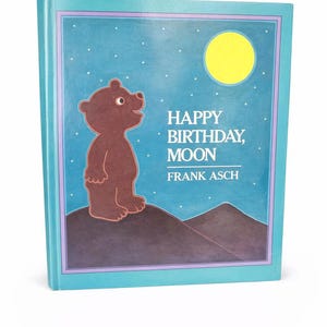 May include: A hardcover children's book titled "Happy Birthday, Moon" by Frank Asch. The cover features a brown bear standing on a hill, with a yellow moon and stars against a blue sky. The book has a teal border.