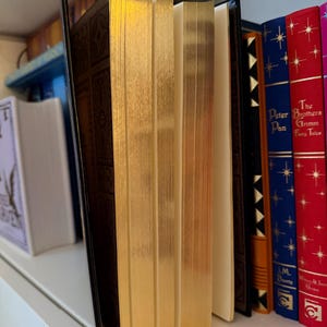 May include: A collection of hardcover books with gold-gilded edges and decorative covers. The books are arranged on a white shelf, with titles visible on the spines, including "Peter Pan" and "Grimm's Fairy Tales."