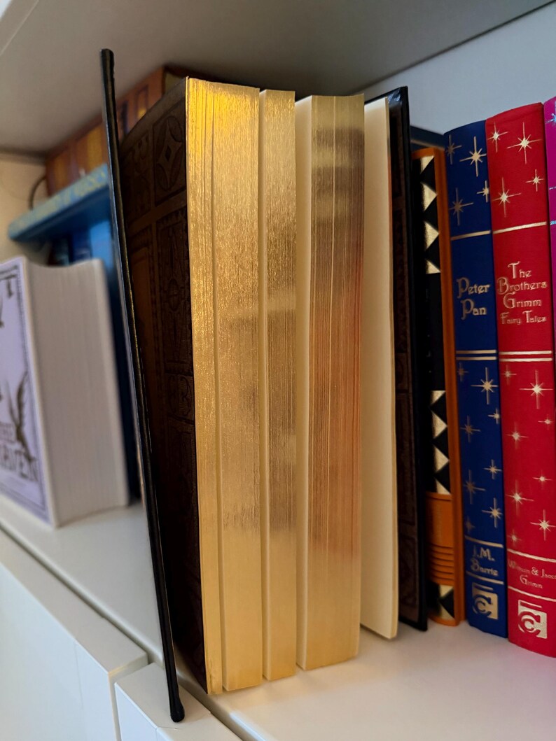 May include: A collection of hardcover books with gold-gilded edges and decorative covers. The books are arranged on a white shelf, with titles visible on the spines, including "Peter Pan" and "Grimm's Fairy Tales."