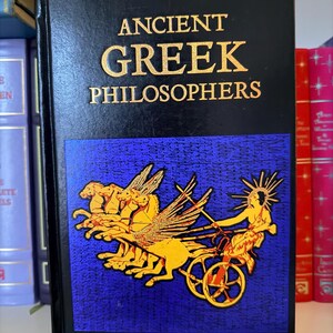 May include: A hardcover book titled "ANCIENT GREEK PHILOSOPHERS" in gold lettering on a black cover. The cover features a blue panel with a gold illustration of a chariot pulled by winged horses. Other books are visible on a shelf.