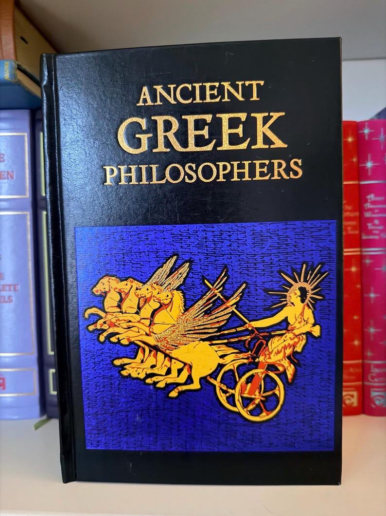 May include: A hardcover book titled "ANCIENT GREEK PHILOSOPHERS" in gold lettering on a black cover. The cover features a blue panel with a gold illustration of a chariot pulled by winged horses. Other books are visible on a shelf.