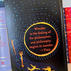May include: A hardcover book with a black cover featuring a quote by Plato: "Wonder is the feeling of the philosopher, and philosophy begins in wonder." The cover has a starry design with gold and blue accents.