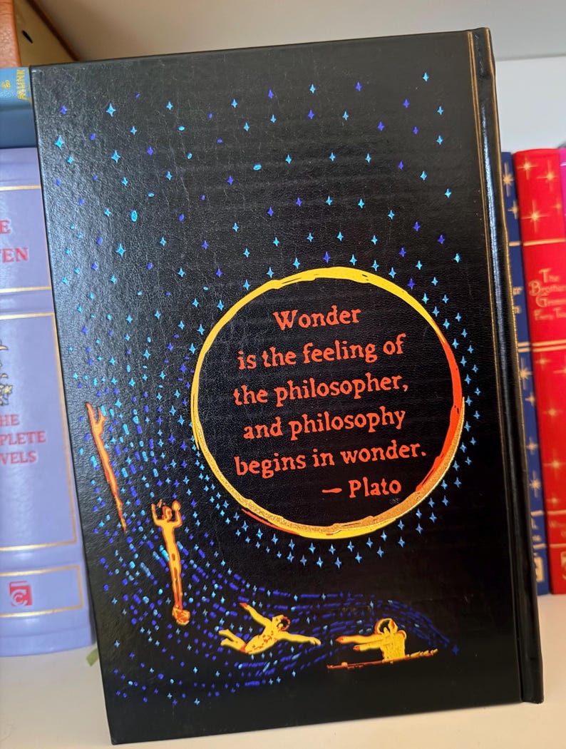 May include: A hardcover book with a black cover featuring a quote by Plato: "Wonder is the feeling of the philosopher, and philosophy begins in wonder." The cover has a starry design with gold and blue accents.