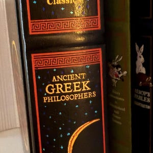 May include: A collection of classic literature books. The primary book is black with gold lettering that reads "Canterbury Classics" and "Ancient Greek Philosophers." The book has a decorative border and a crescent moon design.