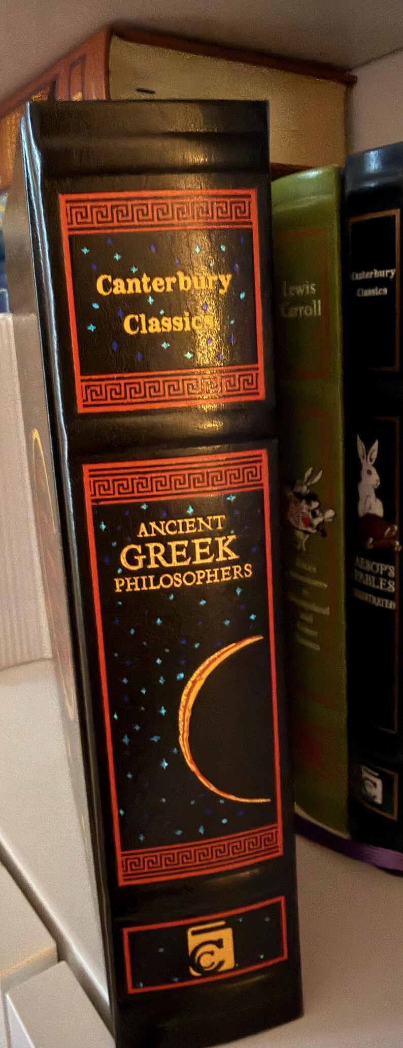 May include: A collection of classic literature books. The primary book is black with gold lettering that reads "Canterbury Classics" and "Ancient Greek Philosophers." The book has a decorative border and a crescent moon design.
