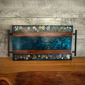 Puede incluir: Una bandeja de servir de resina azul verdosa con un borde de madera y engranajes de metal dorado incrustados en la resina. La bandeja tiene asas de metal negro a los lados.
