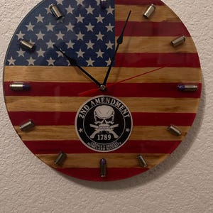 May include: A round wooden clock with an American flag design. The clock face features a black skull and crossbones with the text "2nd Amendment, America's Original Homeland Security, 1789". The clock is decorated with bullet casings around the edge.