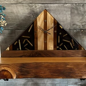 May include: A triangular wooden clock with a unique design. The clock face features alternating dark and light wood panels, with brass bullet casings embedded in the dark sections. The clock hands are black, and the clock sits on a wooden base.