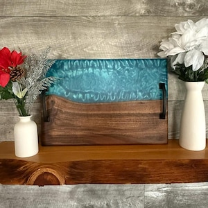 May include: A teal and brown serving tray with black metal handles sits on a wooden shelf. The tray has a teal epoxy resin top with a white swirl design and a brown walnut wood base. Two white vases with flowers are on the shelf.