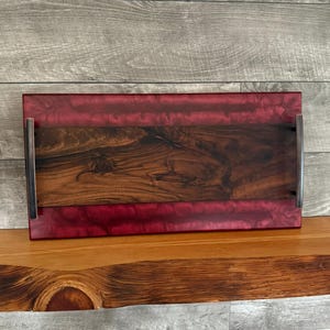 Puede incluir: Una bandeja rectangular de servir hecha de madera y resina. La bandeja tiene una base de madera oscura con una parte superior de resina roja. La bandeja tiene dos asas metálicas en los lados.