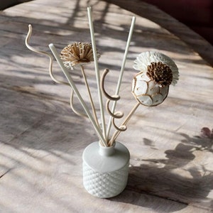 May include: A white ceramic diffuser bottle with a textured surface, filled with natural reed diffusers and decorative dried flowers. The diffuser is on a wooden table with natural light shining through.