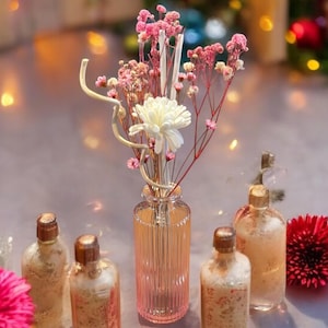 May include: Five glass bottles with gold lids and clear liquid inside. The bottles are arranged in a row on a white surface. A bouquet of pink and white flowers in a clear glass vase is in the centre of the bottles.