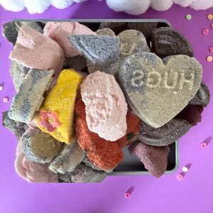 May include: A metal tray filled with colourful, irregularly shaped sweets. The sweets are in shades of pink, yellow, grey, and brown. One sweet is heart-shaped with the word "HUGS" imprinted on it. The background is a solid purple.
