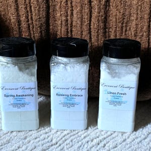 May include: Three white plastic containers with black lids and labels. The labels read "Everscent Boutique" and list different scents: "Spring Awakening", "Relaxing Embrace", and "Linen Fresh". Each container is labelled as a carpet freshener shaker.
