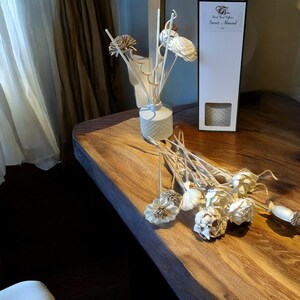 May include: A white ceramic diffuser bottle with a white and gold floral design. The diffuser is filled with reeds and has a sweet almond scent. The diffuser is on a wooden table with a box of reed diffusers in the background.