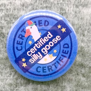 Può includere: Un bottone rotondo blu con le parole "CERTIFIED" e "Certified silly goose" in testo bianco. Il bottone presenta un disegno di un'oca con un cappello. Stelle dorate e un cappello rosso e verde accentuano il design.