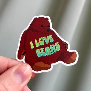 May include: A sticker featuring a cartoon bear in shades of brown and red. The bear has the words "I LOVE BEARS" in green and blue lettering across its chest. The sticker is held by a hand.