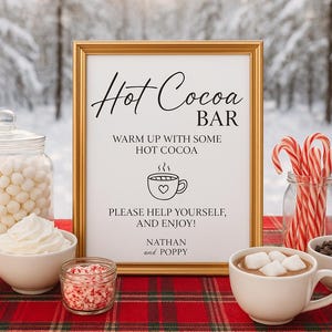 May include: A festive hot cocoa bar setup with a gold-framed sign that reads "Hot Cocoa Bar." Includes marshmallows, whipped cream, chocolate chips, and candy canes. The sign says "Warm up with some hot cocoa. Please help yourself, and enjoy!"