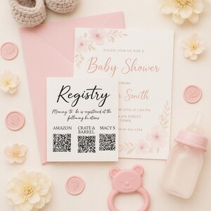 May include: A baby shower invitation and registry card with pink floral accents. The invitation text reads "Please join us for a Baby Shower" and "Registry". The registry card includes three QR codes labeled Amazon, Crate & Barrel, and Macy's. A pink envelope, a baby bottle, and a pink bear-shaped rattle are present.