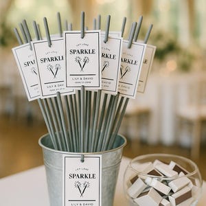 May include: A galvanized metal bucket filled with sparklers, each adorned with a tag that reads "Sparkle" and a wedding date. A small glass bowl holds matchboxes. The scene is set on a white surface.
