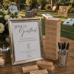 Puede incluir: Un letrero enmarcado de "Libro de visitas Jenga", una torre de bloques de madera Jenga y bloques inscritos con buenos deseos. Los marcadores negros están en un soporte de vidrio. La escena está ambientada al aire libre.