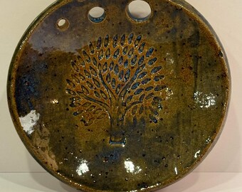 May include: A round, ceramic dish with a tree design in brown and blue glaze. The dish has three holes around the rim.