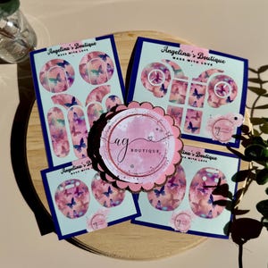 Könnte beinhalten: Vier Blätter mit pinkfarbenen und weißen Schmetterlingsaufklebern mit dem Text "Angelina's Boutique Made With Love". Die Aufkleber sind auf einer Holzoberfläche mit einem pinkfarbenen und weißen Schmetterlingsaufkleber in der Mitte angeordnet.