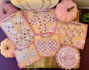 May include: A collection of Halloween-themed stickers and earrings featuring ghosts and pumpkins in pink and white. The stickers are displayed on pink cards with the text "Angelina's Boutique" and "Made with Love". Pink pumpkins are also in the image.