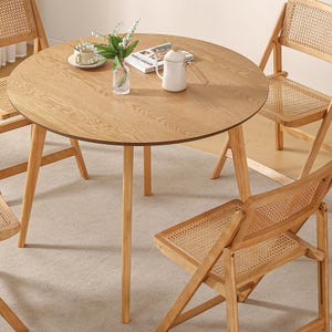 Könnte beinhalten: Ein runder Holztisch mit vier Stühlen. Die Stühle sind aus Holz und haben geflochtene Korbsitze. Der Tisch hat eine helle Holzoptik und ist von einem hellbeigen Teppich umgeben.