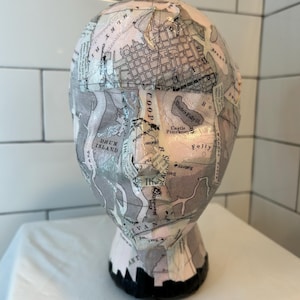 Può includere: Una testa di manichino in ceramica bianca con una base nera, decorata con un collage di mappe e testi antichi. Le mappe includono dettagli di "Sturteis Folly Id", "Castle Pinckney", "Folly", "Drum Island" e "Coop".