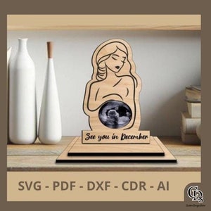 Pode incluir: Corte de madeira de uma mulher grávida segurando uma imagem de ultrassom em sua barriga. O corte está sobre uma base de madeira com o texto "See you in December".