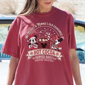 May include: A maroon t-shirt featuring Mickey and Minnie Mouse with the text "Mickey & Minnie's Old Fashioned Hot Cocoa Served Daily". The shirt is a relaxed fit and has short sleeves. The background includes a white convertible car.